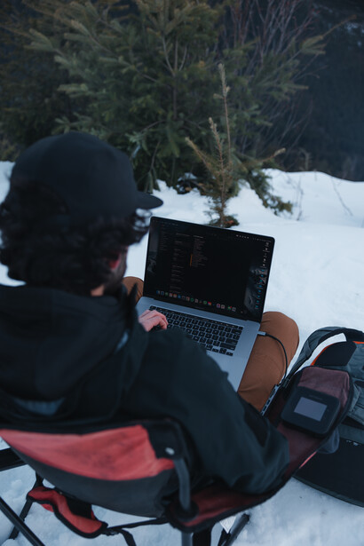 Photographer working on laptop in snowy outdoor setting with evergreen tree and camera equipment visible.