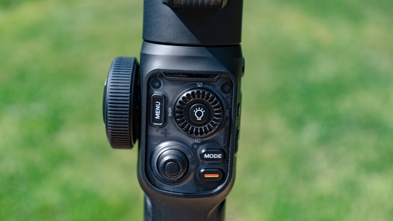 Close-up of a camera flash unit's control panel with illuminated dial, mode button, and adjustment knob against blurred green background.