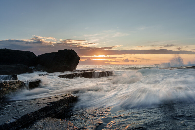 Waves crash against rocky shoreline during golden hour sunset with dramatic cloudy sky.
