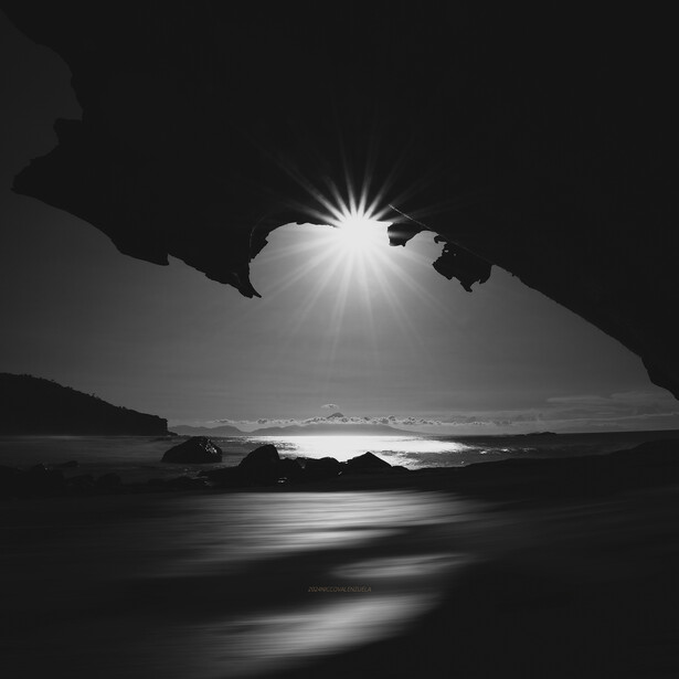 Sunlight bursts through a cave opening onto a beach with flowing water below.
