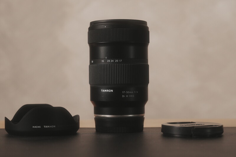 Tamron lens with hood and accessories arranged on a dark surface against a neutral background.