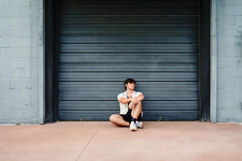 Person in white t-shirt sitting against closed blue metal garage door on concrete ground.