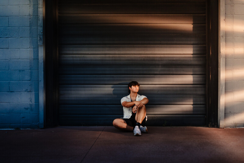Shirtless man sitting cross-legged against a dark corrugated metal door, arms crossed, with geometric shadow patterns from sunlight.