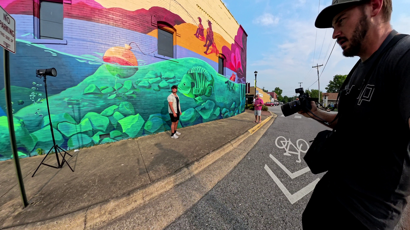 Photographer with camera on tripod documenting large-scale colorful mural on building wall.