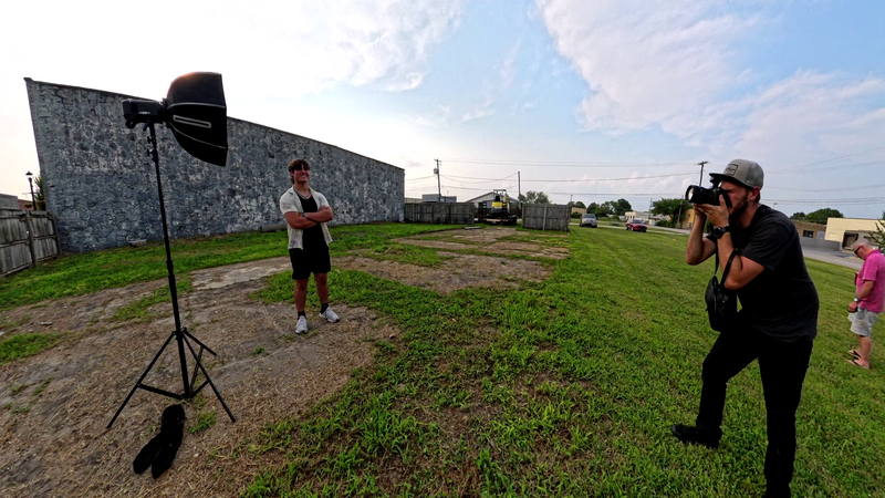 Photographer with camera on tripod shooting a subject standing in front of a large gray backdrop in an open field.
