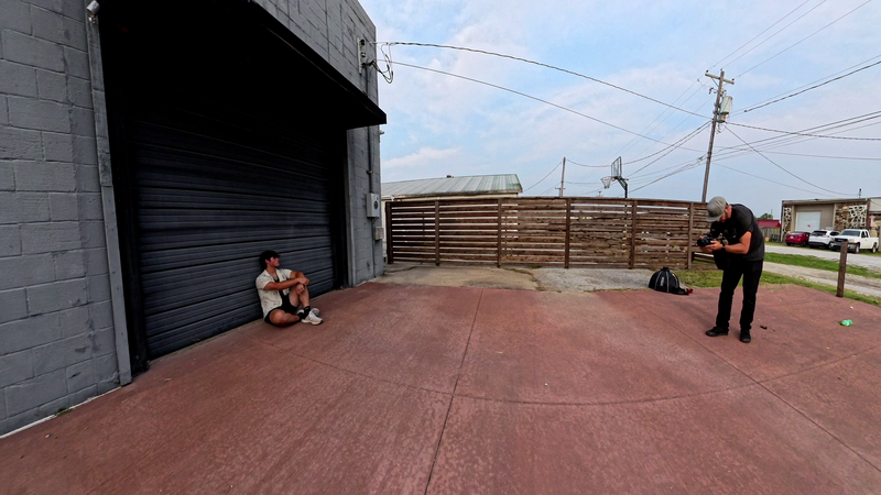 Two people positioned in an empty paved yard next to a gray building with a dark rolling door, preparing for or during a photo shoot.