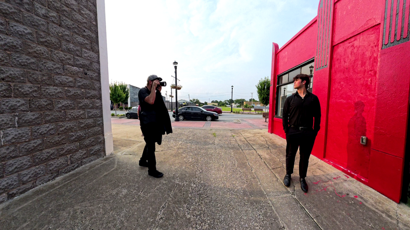 Two people in dark clothing standing in an empty parking area between a gray textured wall on the left and a bright red metal structure on the right.