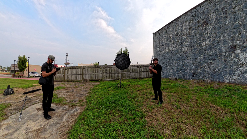 Two people in black clothing hold guide wires on either side of a young tree in an open field next to a stone building.
