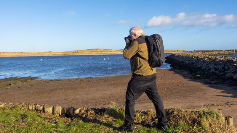 Photographer with backpack holding camera to face while standing on beach looking toward water.