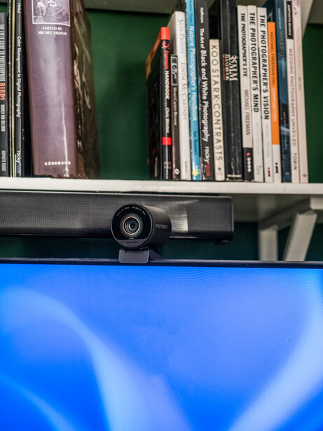 Webcam mounted on top of a monitor above a shelf of books and media.