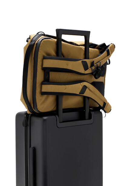 Tan canvas camera backpack with black straps mounted on top of a black rolling suitcase.