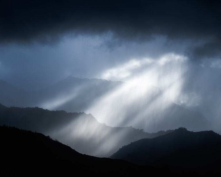 Dramatic crepuscular rays streaming through dark storm clouds above silhouetted mountain ridges.