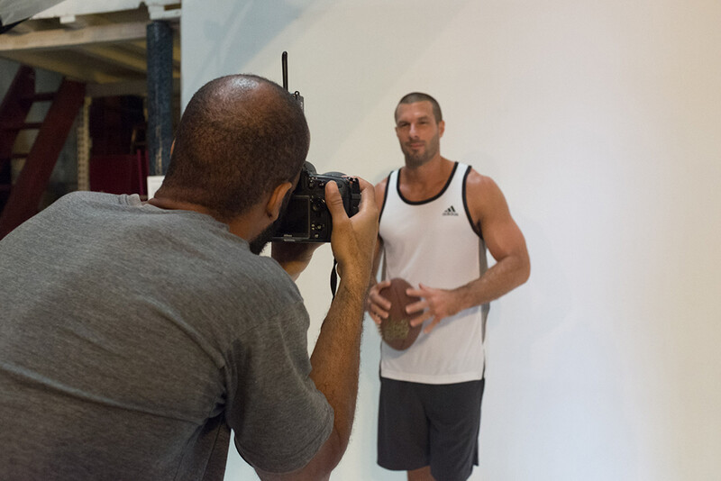 Photographer holding a camera takes a portrait of a man in a white tank top against a white wall.