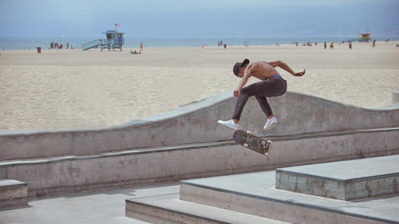 Shirtless skateboarder performing a trick on concrete ledges with beach and ocean in background.