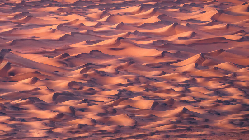 Desert sand dunes with warm golden-orange lighting and deep shadow detail across undulating ridges.
