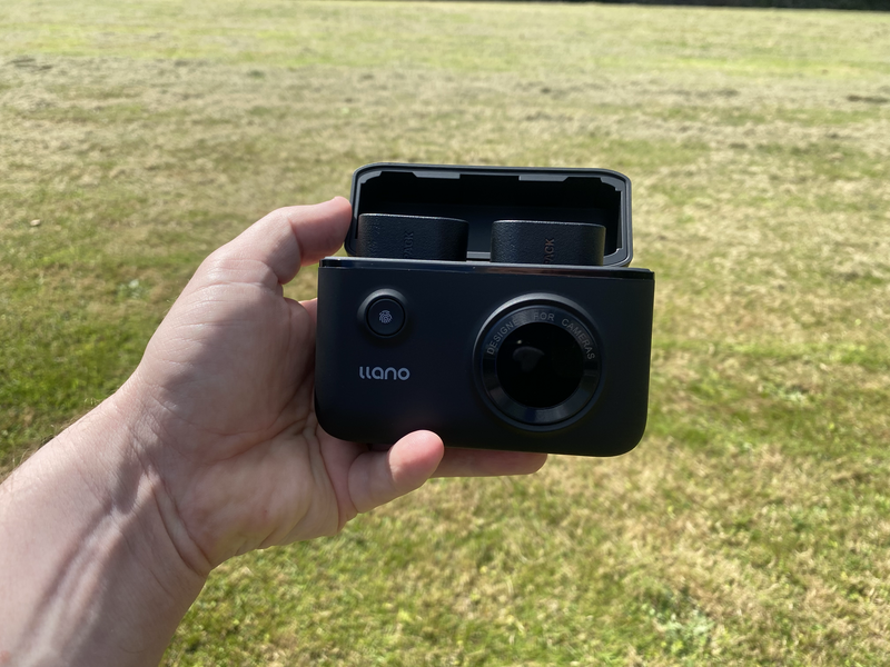 Hand holding a black Llano instant camera with open film compartment against a grass background.