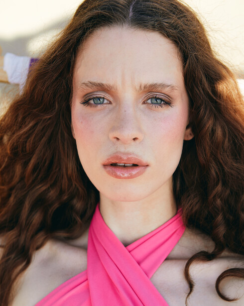 Portrait of a woman with long wavy brown hair and blue eyes wearing a bright pink crossed neckline garment.