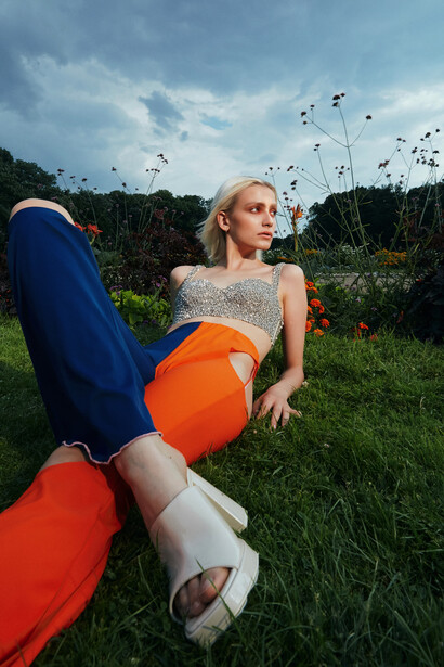 Woman in grass holding orange balloon, wearing blue pants and gray top, looking away from camera.