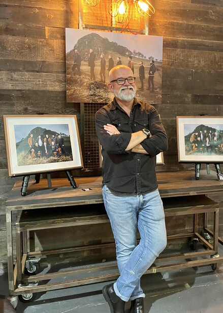 Artist standing with arms crossed in front of displayed landscape paintings in a studio setting.