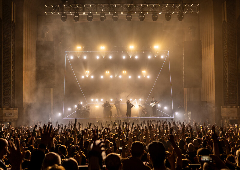 Large crowd with raised hands facing an illuminated stage with geometric frame structure and warm overhead lighting.