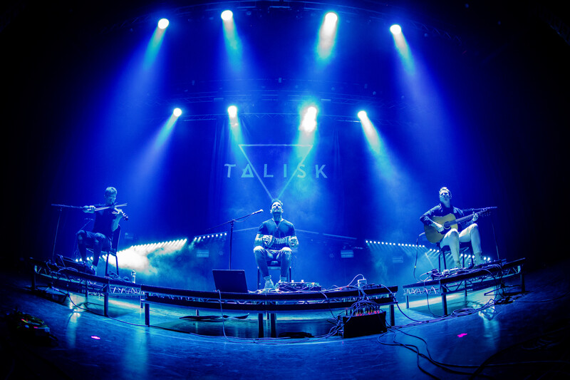 Concert stage with band performing under blue spotlights and atmospheric lighting effects.