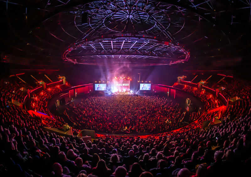 Large concert venue filled with thousands of spectators, illuminated by red and purple stage lighting and overhead geometric light fixtures.