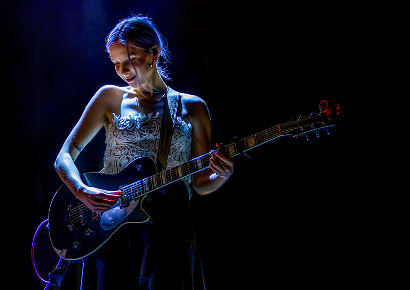 Female guitarist performing on stage under blue stage lighting while playing electric guitar.
