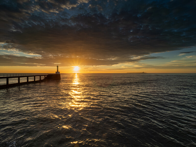 Sun setting over calm ocean waters with a pier extending into the sea on the left side.