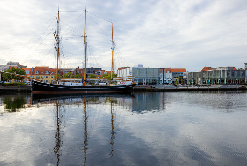 Traditional sailing vessel with three masts moored in calm water, reflecting perfectly in mirror-like surface.