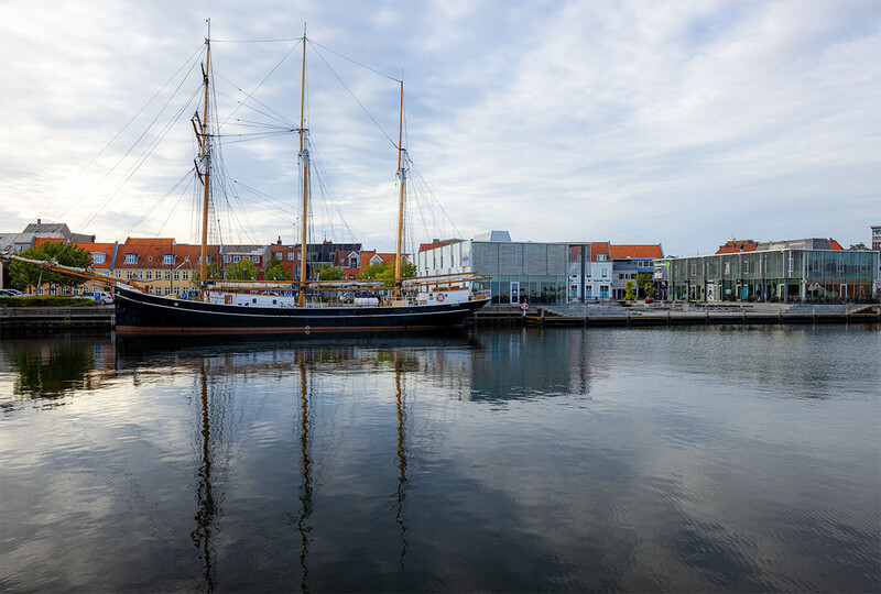 Traditional sailing vessel with three masts reflected in still water alongside historic waterfront buildings.