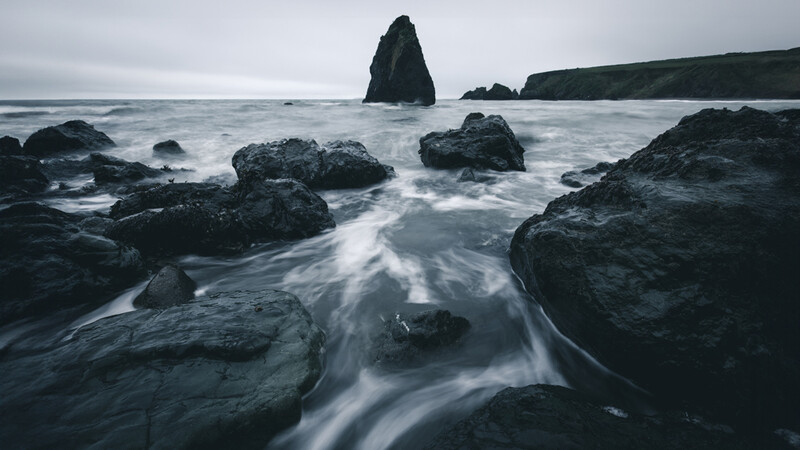 Dramatic coastal seascape with tall dark rock formations and smooth water flowing around boulders.