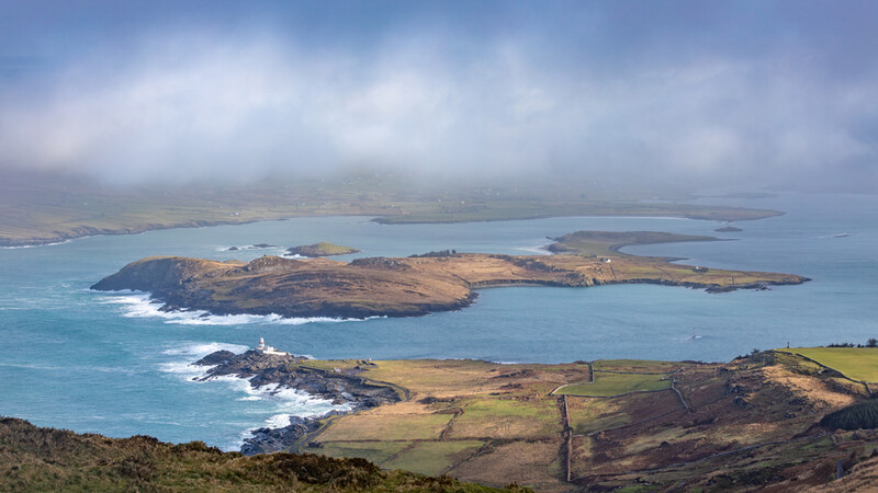 Aerial view of volcanic islands with brown grassland and turquoise ocean waters under cloudy sky.