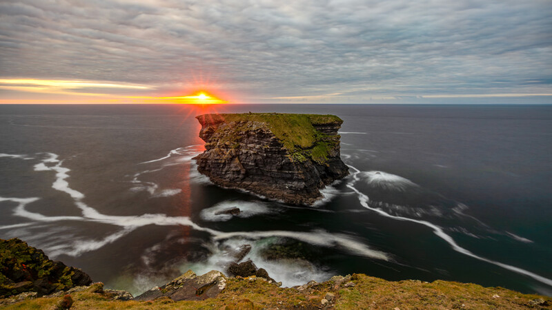 Rocky sea stack surrounded by crashing waves at sunset, viewed from a coastal cliff with golden hour light breaking through cloudy sky.