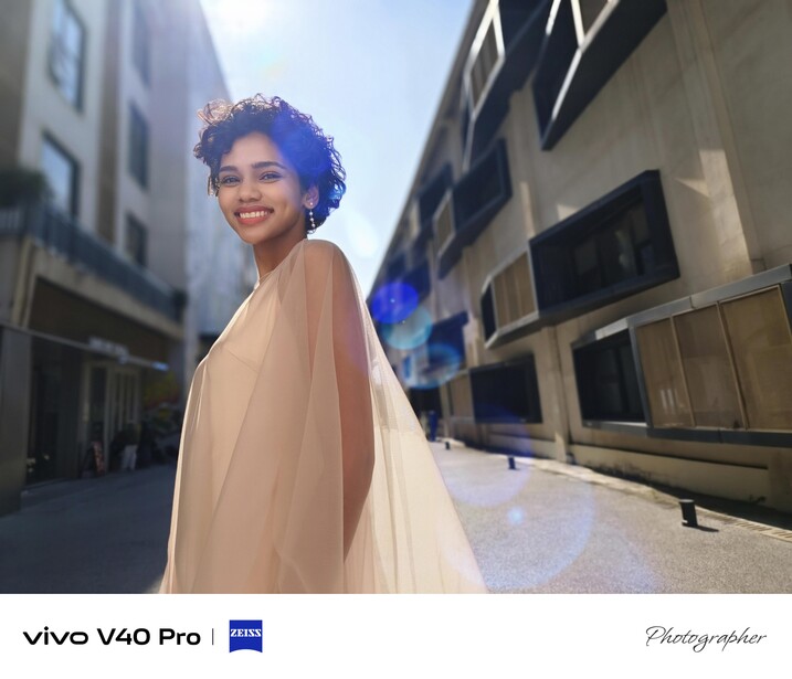 Woman in flowing beige cape smiling at camera in urban alleyway between modern buildings.