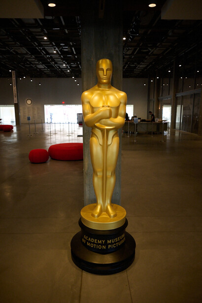 Large golden Oscar statuette on black pedestal in modern gallery space with industrial ceiling and red seating.