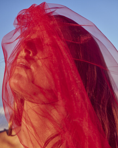 Person wearing vibrant red sheer fabric draped over their head and shoulders against a clear blue sky.