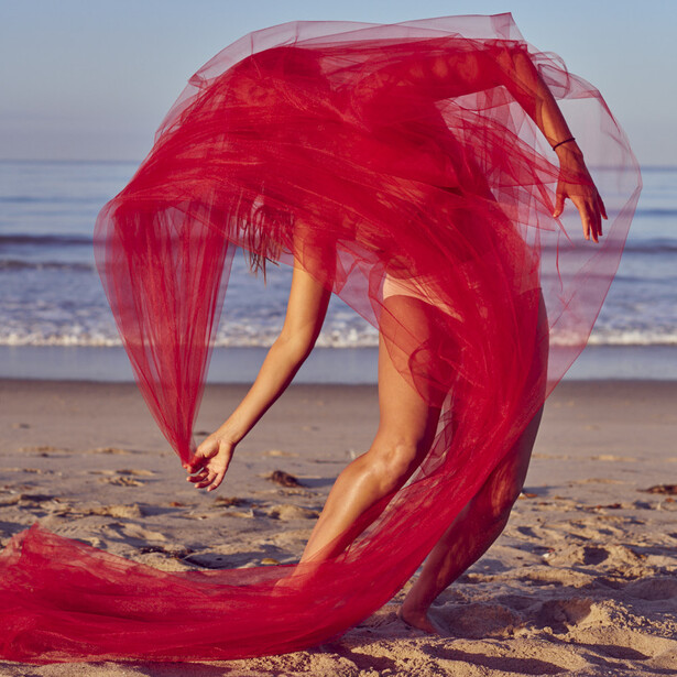 Woman on beach holding flowing red fabric that curves around her body.