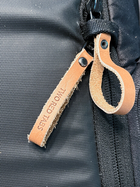 Leather camera gear tags and carabiner clips attached to a dark gray camera bag zipper.