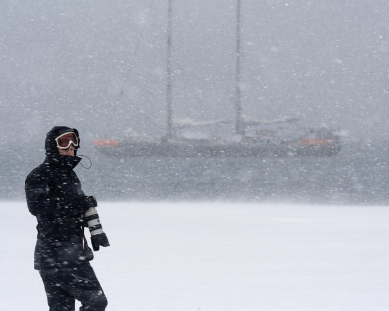 Snowboarder in black jacket and goggles standing on snowy slope during heavy snowfall.