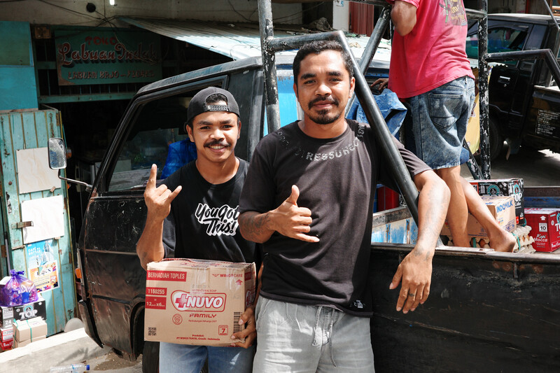 Two men posing with thumbs up in front of a pickup truck loaded with boxes and merchandise.