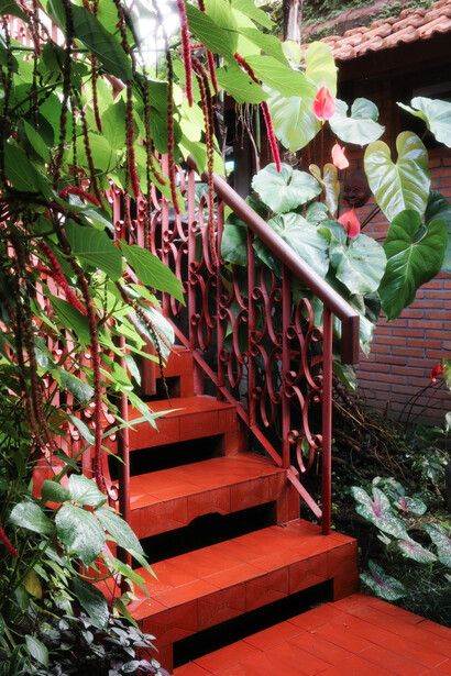 Vibrant red staircase with ornate metal railing surrounded by lush green foliage and flowering plants.