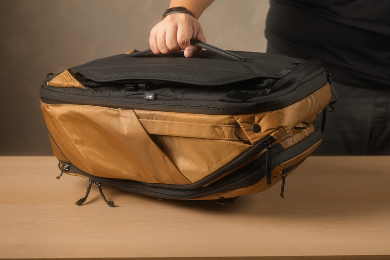 Person opening a tan and black camera bag with multiple compartments on a wooden surface.