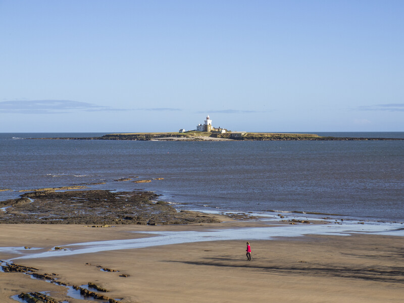 Solitary figure in red walking on wet sand beach with distant lighthouse across the water.
