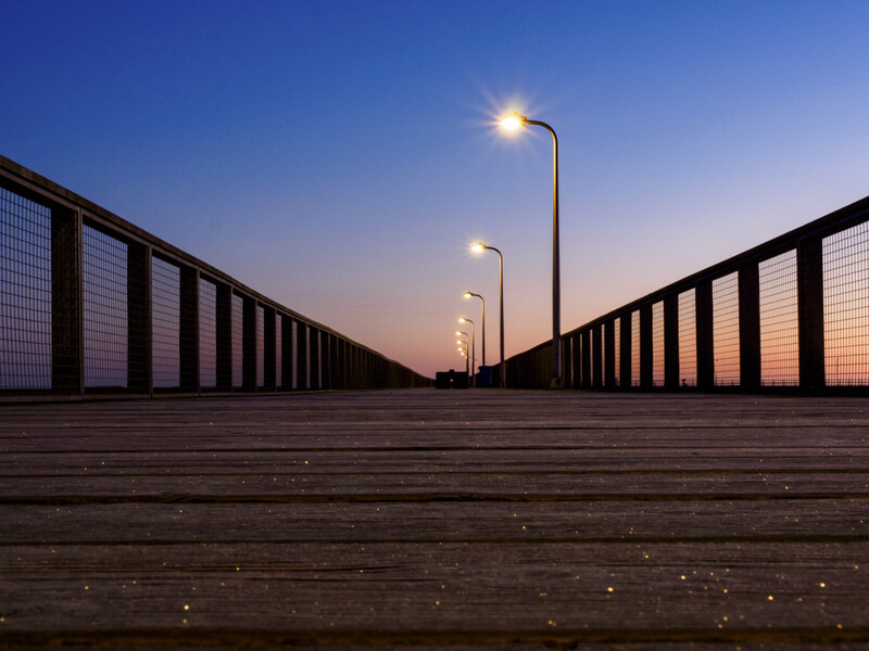 Wooden boardwalk with metal railings extending toward the horizon at dusk, lit by street lamps.