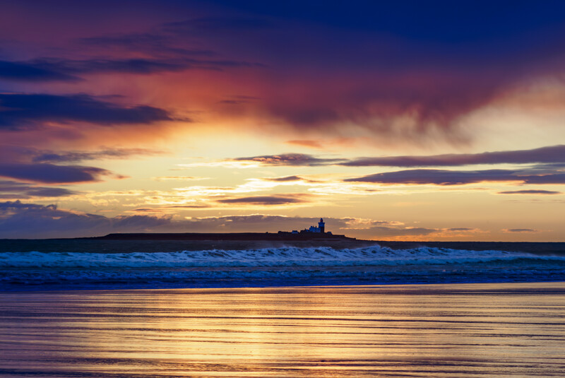Dramatic sunset sky with golden and purple tones reflected on wet beach sand and ocean waves.
