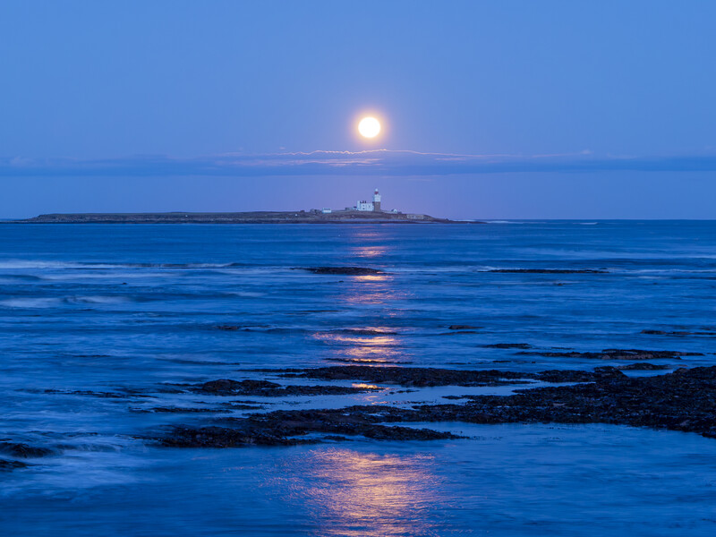 Full moon rising above the ocean horizon with rocky coastline in foreground at twilight.
