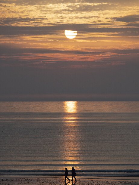 Two figures silhouetted against the ocean during golden hour sunset with sun reflecting on calm water.