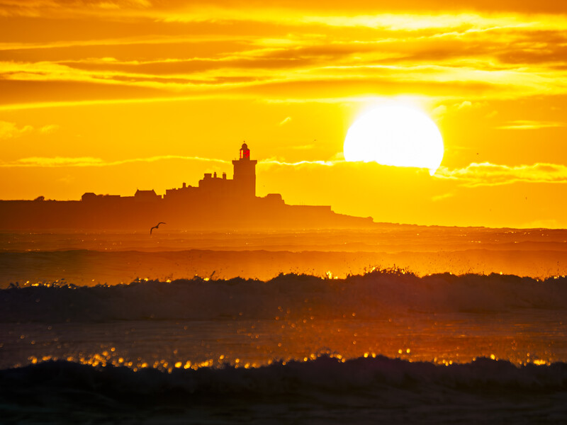 Silhouetted coastal town with church tower against massive golden sunset sun.