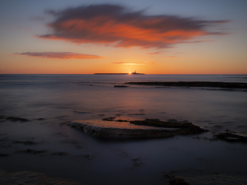 Sunset over a rocky coastline with orange and red clouds reflecting on calm water.