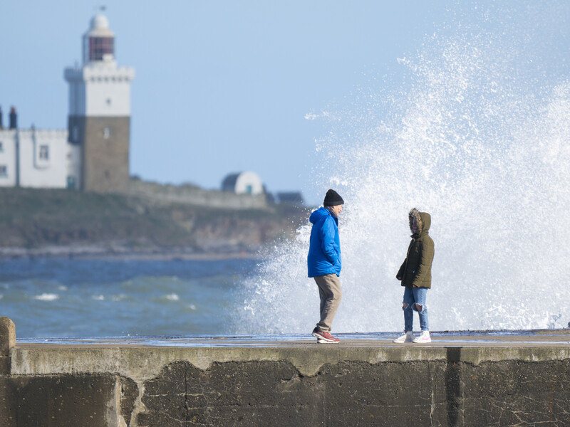 Two children standing on a concrete pier as a large wave splashes behind them, with a lighthouse visible on the shoreline.